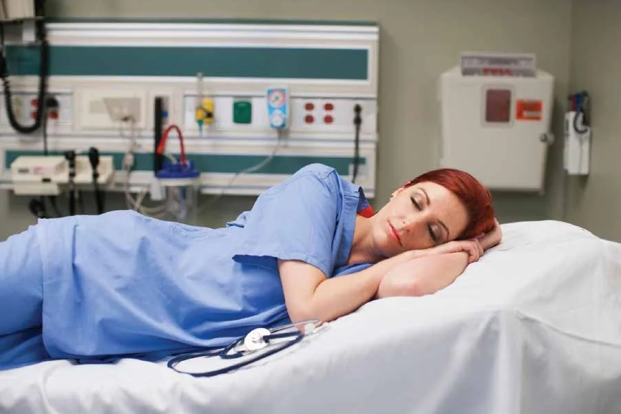 Exhausted hospital nurse resting on a patient bed during a break after a long shift, illustrating the effects of fatigue and nurse staffing shortages