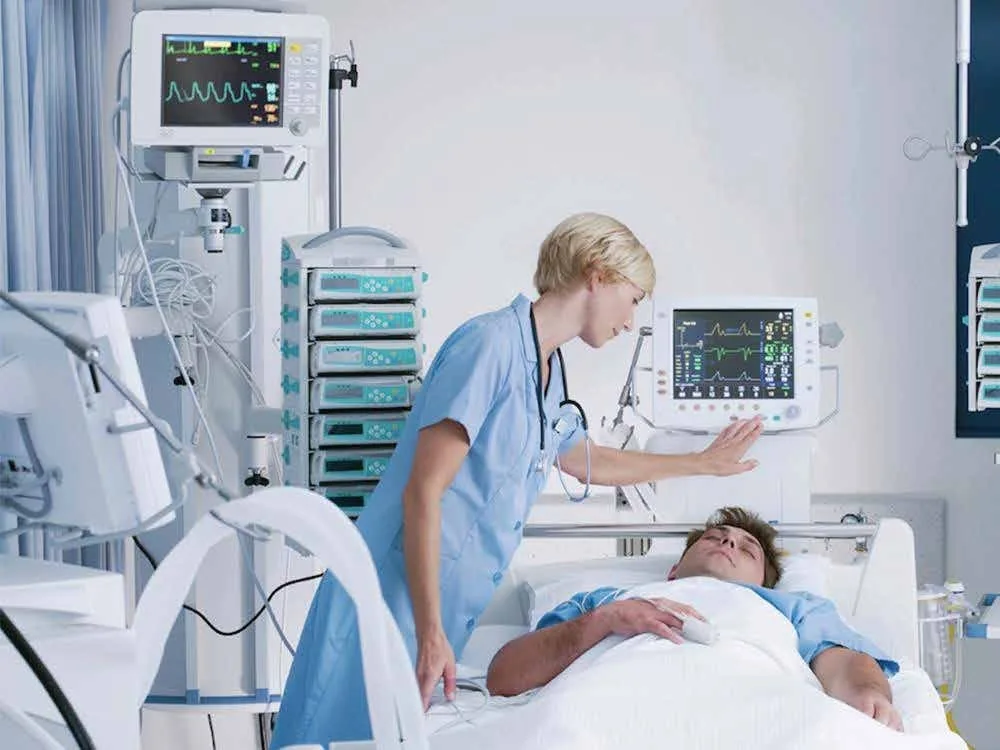 ICU nurse monitoring a patient's vital signs using bedside monitoring equipment in a hospital intensive care unit.