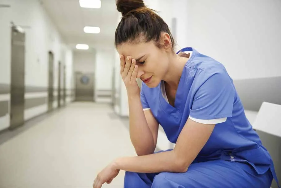 Exhausted hospital nurse sitting in a hallway during a break, illustrating nurse burnout caused by heavy workloads and staffing shortages